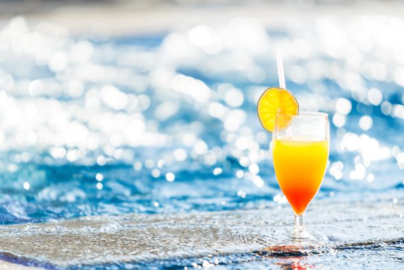 Ein oranger Cocktail steht am Strand vor glitzerndem Meer