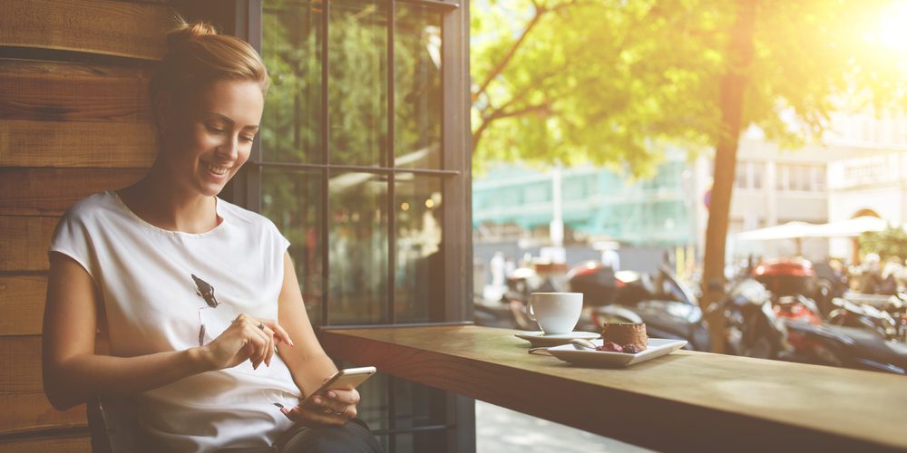 Lächelnde Frau sitzt bei offenen Fenster am Handy - Tipps für mehr Me-Time