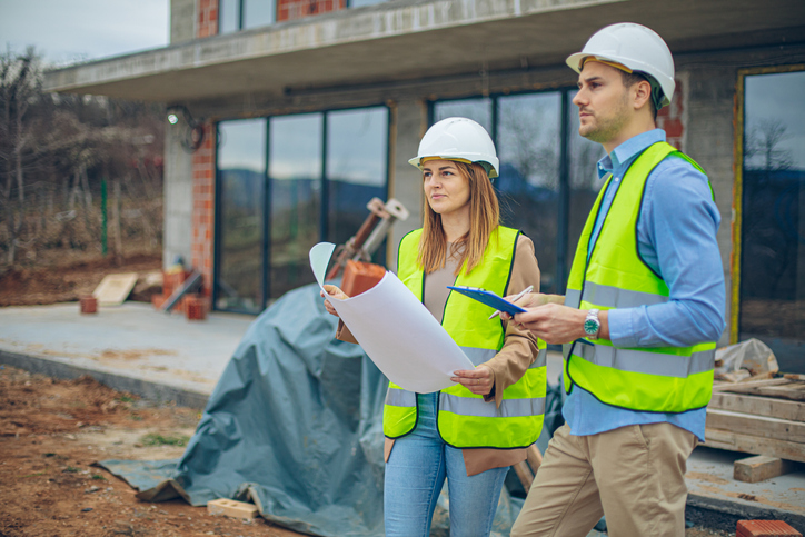 Ein Mann und eine Frau stehen mit Warnweste, Schutzhelm und Plan vor einem Einfamilienhaus. Planung ist auch in der Asset Allocation essentiell.