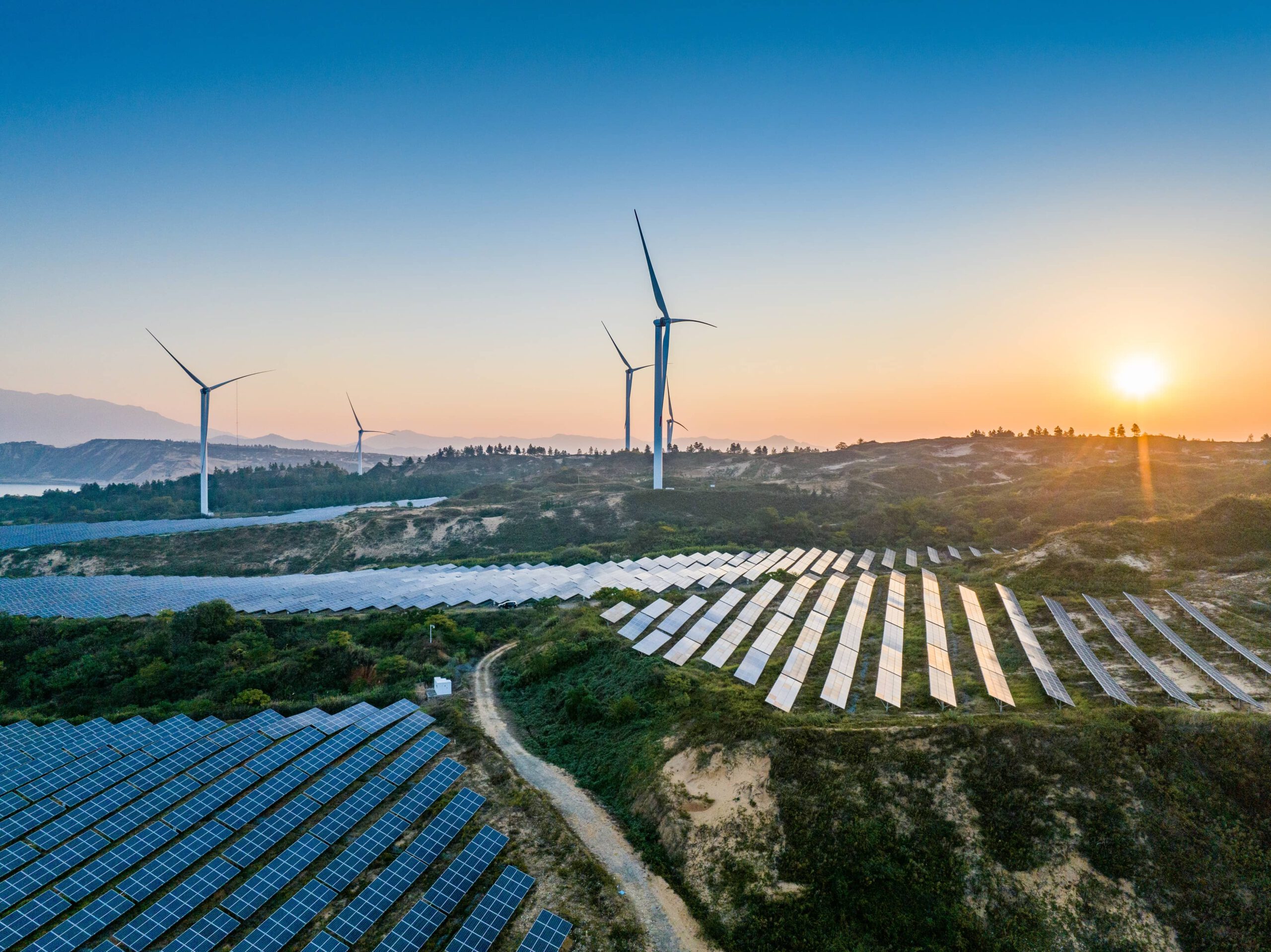 Das Bild zeigt einen Weitblick in die aufgehende Sonne. Man sieht Windräder und Photovoltaikanlagen, wichtige Bereiche erneuerbarer Energie.