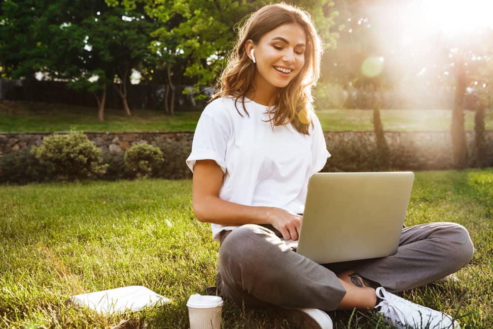 Junge Frau sitzt in der Natur und schaut in ihren Laptop.